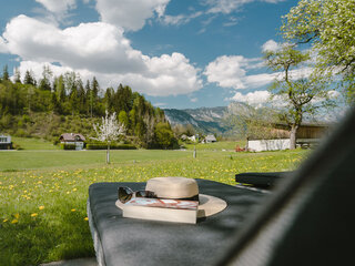 Ein Buch und ein Hut liegen am Liegestuhl. Rund herum ist eine Wiese und im Hintergrund sieht man Berge und Häuser.